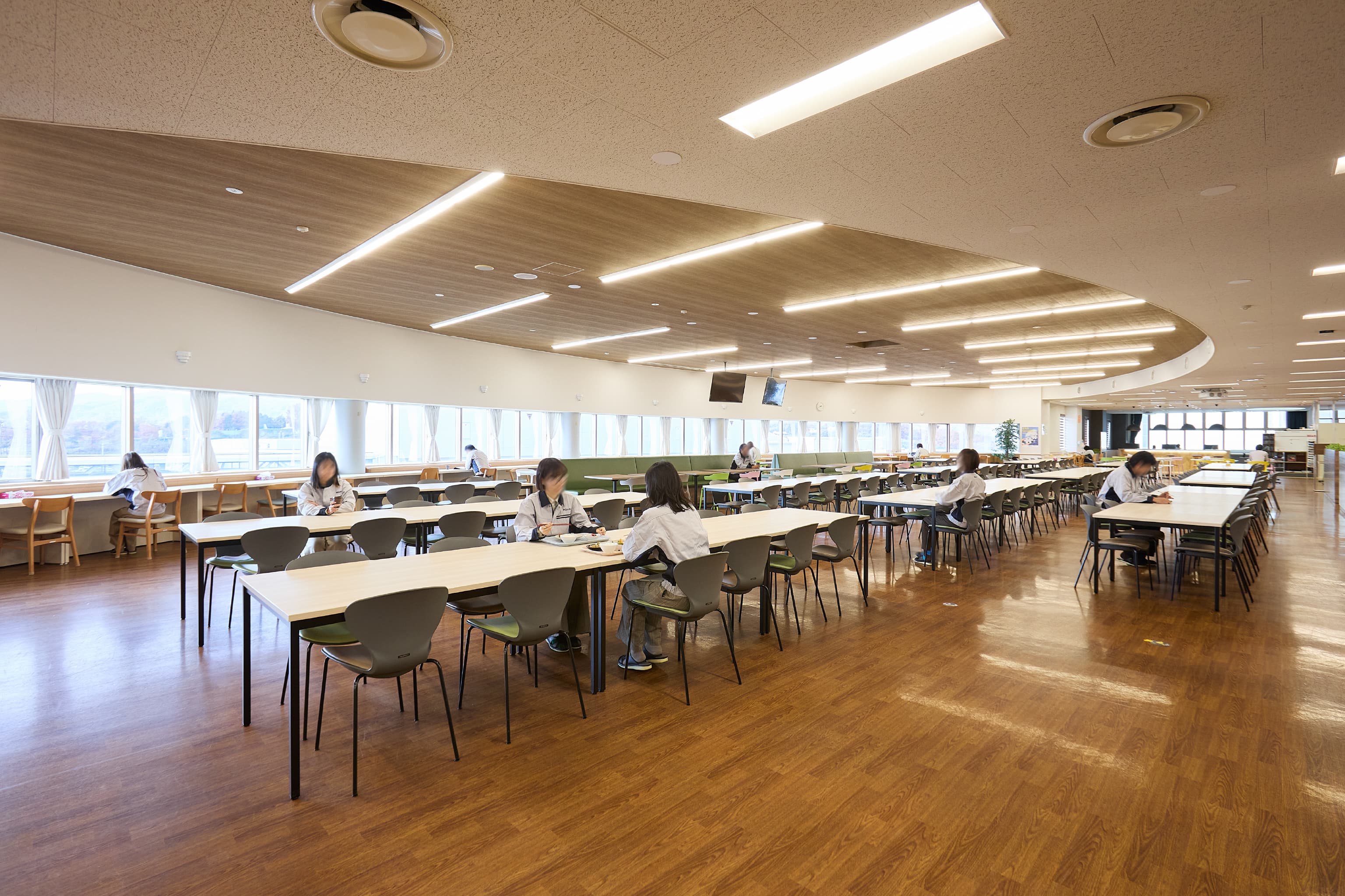 日東電工株式会社 様の事例写真／[3F 社員食堂]もとからある床材に呼応するように天井も木目張りとし、「明るくあたたかみのある空間になった」と好評です。カーブに沿って並ぶ、光が差し込む窓際席は、集中したいときや自分の時間を過ごしたいときに人気です。