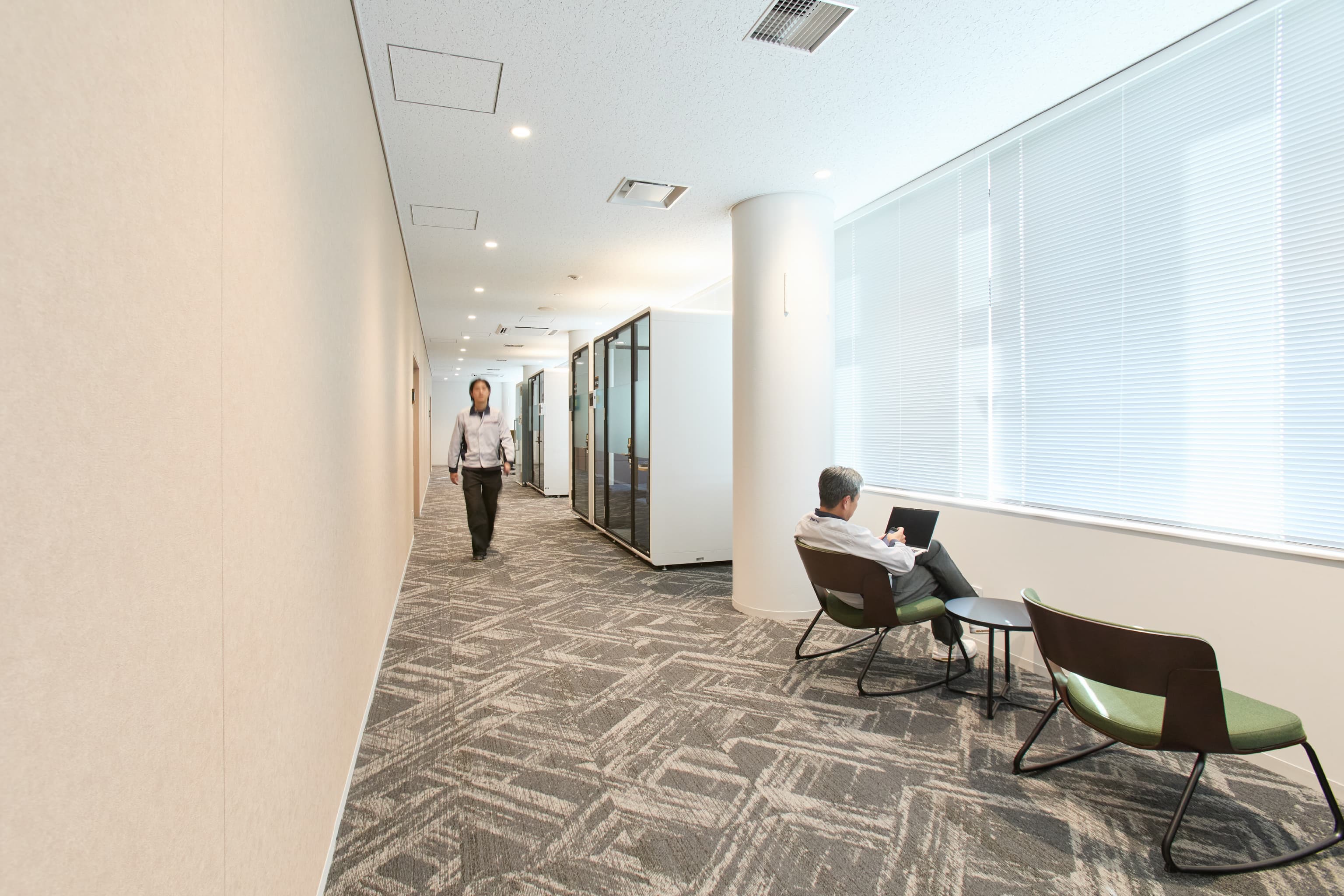 日東電工株式会社 様の事例写真／[6F 通路]通路脇には、ロッキングチェアがさりげなく配置され、パソコン操作をしたり、会議を待ったりする人の姿が見られます。鳥がさえずる心地よい環境音が流れ、ふと耳を傾けるだけでもリラックスできます。